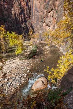 Two unidentifiable hikers walking through the Virgin River on the Narrows path at Zion National Park, Utah.