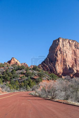 Kolob Terrace Road paved in red rock leading into tall sandstone mountains that have been eroded over time and a blue sky background in Zion National Park, Utah.
