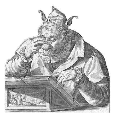 Foto de Un hombre con una capucha de bufón en la cabeza y gafas en la nariz hinchada se sienta frente a un atril en el que yace un libro - abierto, pero con la cubierta hacia arriba. - Imagen libre de derechos