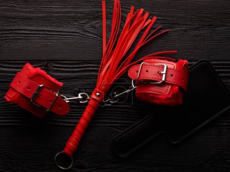 Photo for Bright red handcuffs, red whip and black paddle over black wooden background - Royalty Free Image