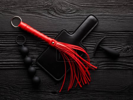 Bright red handcuffs and red whip over black wooden background