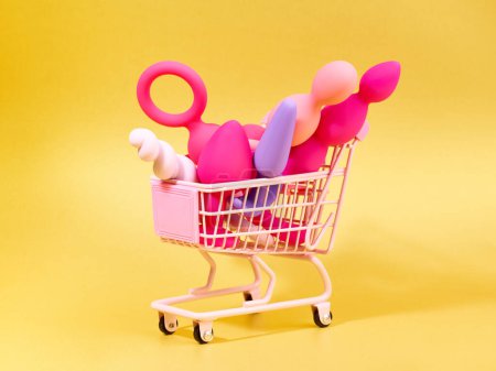 Téléchargez les photos : Anal plugs and dildo sex toys in shopping basket over yellow backdrop - en image libre de droit