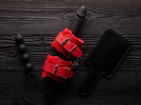 Knallrote Handschellen und rote Peitsche über schwarzem Holzhintergrund