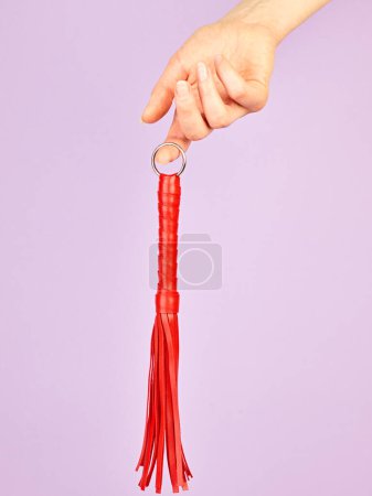 Foto de Látigo rojo para juegos de rol para adultos en la mano de la mujer sobre fondo violeta - Imagen libre de derechos