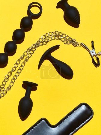 Téléchargez les photos : Black sex toys dildo, anal plugs and clamps over yellow paper background - en image libre de droit