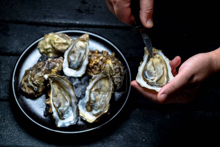 Photo for Oyster in hands opens with a knife on the background of a dish with oysters - Royalty Free Image