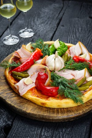 Foto de Pizza con jamón, pepinos en escabeche, ajo, pimientos rojos y queso en una mesa de madera. - Imagen libre de derechos