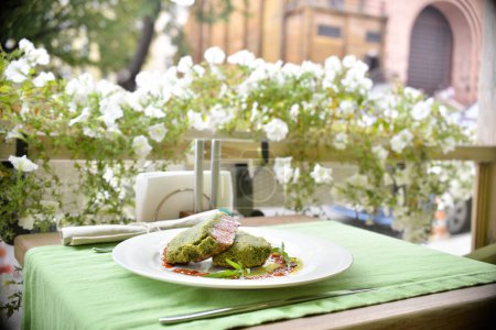 Foto de Un primer plano de carne empanada verde en un plato blanco con salsa en la terraza con flores - Imagen libre de derechos
