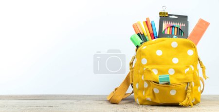 Photo for Yellow backpack with white polka dots with different colorful stationery on table. White background. Back to school. Banner design - Royalty Free Image