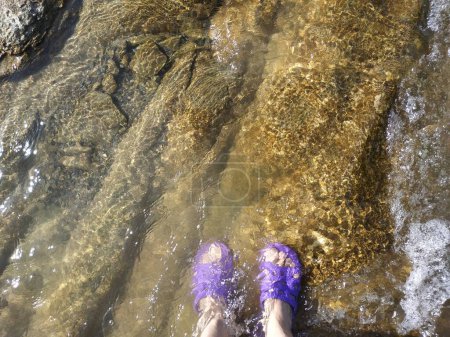 Téléchargez les photos : Pieds féminins en tongs, pantoufles immergées dans l'eau près du rivage, sur pierre par temps ensoleillé - en image libre de droit