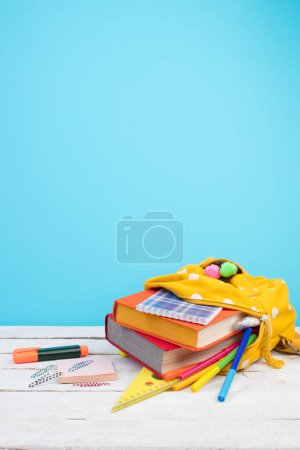 Foto de Mochila amarilla con lunares blancos con diferentes artículos de papelería de colores en la mesa. Fondo azul. De vuelta a la escuela. Diseño de banner - Imagen libre de derechos