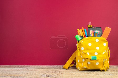 Backpack with different colorful stationery on table. Burgundy background. Back to school