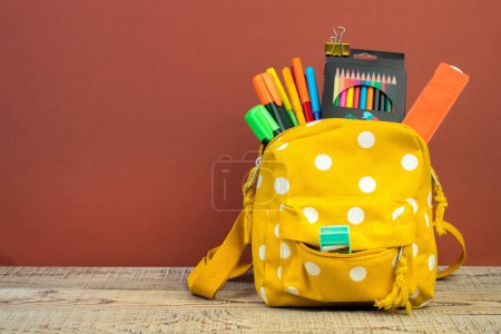 Photo for Backpack with different colorful stationery on table. Brown background. Back to school - Royalty Free Image