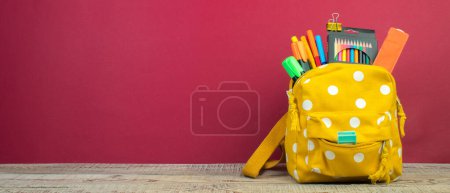 Foto de Mochila con diferentes artículos de papelería de colores en la mesa. Fondo borgoña. Regreso a la escuela - Imagen libre de derechos