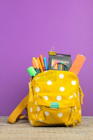Photo for Backpack with different colorful stationery on table. Purple background. Back to school. - Royalty Free Image