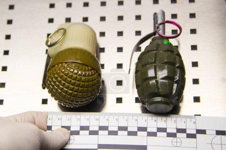Téléchargez les photos : Deux grenades à fragmentation portatives en métal munies d'une règle médico-légale et d'une main experte en gants de caoutchouc. - en image libre de droit