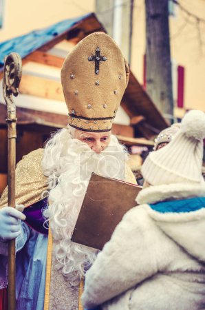 Saint Nicolas rencontre un enfant au Marché de Noël pendant que la famille attend en arrière-plan
