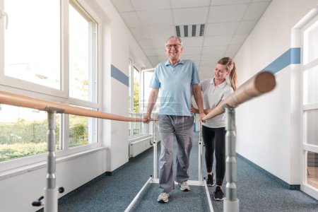 Téléchargez les photos : Patiente aînée et physiothérapeute dans des exercices de réadaptation à pied, elle l'aide le long des barreaux - en image libre de droit