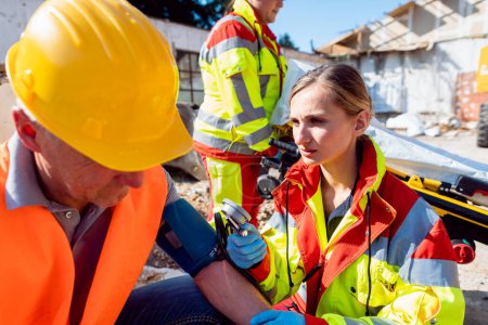 Téléchargez les photos : Médecin paramédical mesurant la pression artérielle et parlant à l'homme après un accident sur le chantier - en image libre de droit