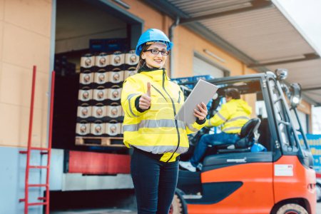 Photo for Woman in industrial gear standing in front of a forklift transporting goods from a warehouse - Royalty Free Image