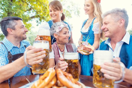 Téléchargez les photos : Groupe d'amis dans le jardin de la bière toastant à la caméra portant tracht bavarois - en image libre de droit