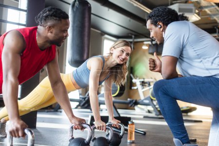 Photo for Diversity woman and man in modern gym doing push-ups with kettle bells under supervision of coach - Royalty Free Image