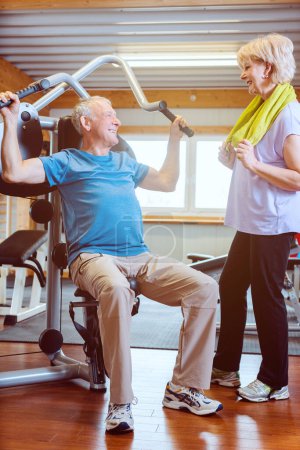 Téléchargez les photos : Heureux homme âgé dans la salle de gym avec sa femme sur les machines d'exercice - en image libre de droit