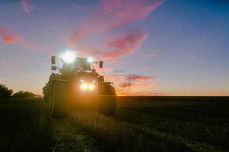 Téléchargez les photos : Fin d'une journée de récolte sur un champ de céréales en été avec tracteur avec la dernière lumière au crépuscule - en image libre de droit