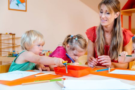 Children drawing in playgroup of nursery school with the teacher helping