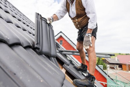 roofer in traditional German clothing of a craftsman working with roof tiles on the roof.