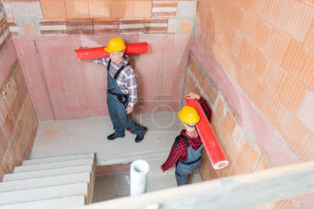 Téléchargez les photos : Vue aérienne de deux ouvriers de la construction portant des tuyaux rouges sur leur escalier marchant à l'épaule - en image libre de droit