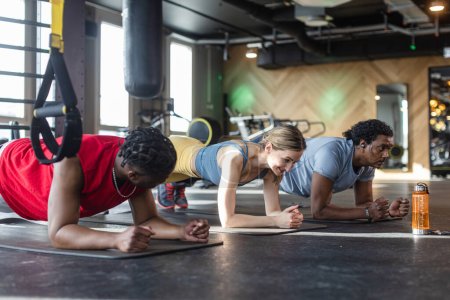 Foto de Tres personas, dos hombres negros y una mujer caucásica, haciendo tablones y abdominales de entrenamiento en un gimnasio moderno - Imagen libre de derechos