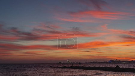 Photo for In the Bay of Palma, a sunset casts reddish hues. The sea meets a jetty with silhouetted fishermen. Far off, a cruise ship's lights dot the horizon - Royalty Free Image
