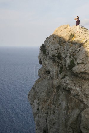 Téléchargez les photos : Au-dessus de la mer bleue sans fin, une figure solitaire se dresse sur une falaise accidentée, immergée dans l'acte de capturer l'horizon - en image libre de droit
