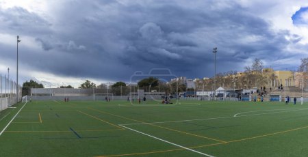 A panoramic view of a soccer match in progress with players and spectators under a dramatic overcast sky.