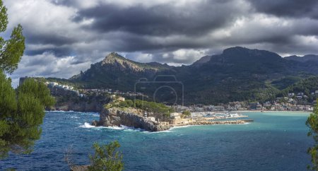 Storm clouds gather over the tranquil blue waters of Soller Port, framed by lush greenery and the rugged Tramuntana mountains in Mallorca.