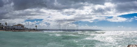 Panoramic view of Portixol's serene coastline, with turbulent waves meeting the shore beneath a stormy sky