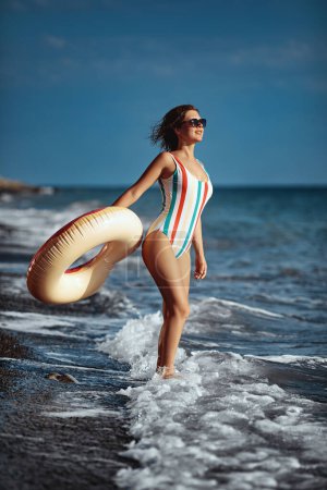 Photo for Smiling young woman in a swimsuit and sunglasses standing on a beach with an inflatable ring and looking at the see. - Royalty Free Image