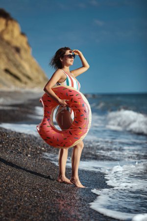 Téléchargez les photos : Jeune femme bronzée en maillot de bain et lunettes de soleil debout sur une plage avec un anneau gonflable et regardant la mer. - en image libre de droit