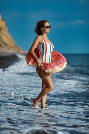 Photo for Tanned young woman in a swimsuit and sunglasses standing on a beach with an inflatable donut ring and looking at the see. - Royalty Free Image