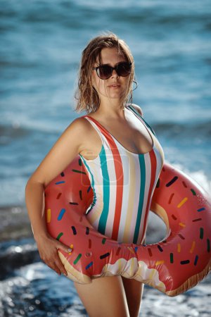 Photo for Tanned young woman in a swimsuit and sunglasses standing on a beach with an inflatable donut ring and enjoying sunbathing. - Royalty Free Image