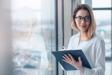 Photo for Young woman standing at the panoramic window of a modern office with a folder of documents and a pen. - Royalty Free Image