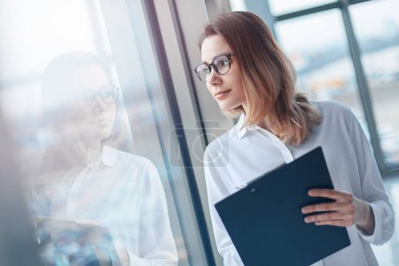 Photo for Young woman standing at the panoramic window of a modern office with a folder of documents. - Royalty Free Image