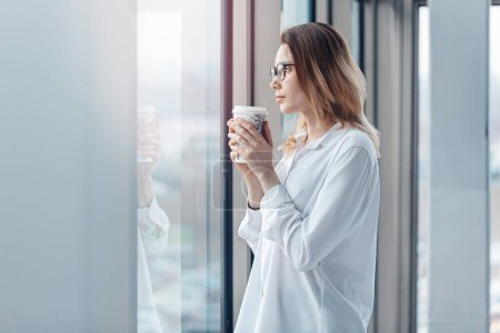 Téléchargez les photos : Jeune femme debout à la fenêtre d'un bureau moderne et tenant une tasse de café. - en image libre de droit