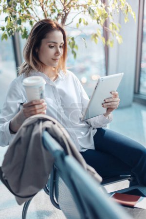 Foto de Mujer viajera joven con una tableta y café esperando un vuelo en la sala de salidas en el aeropuerto. - Imagen libre de derechos