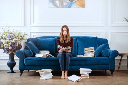 Photo for Young woman reads among stacks of books in a spacious living room. - Royalty Free Image