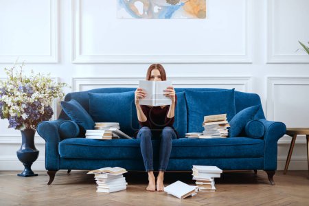 Photo for Young woman covers her face with a book in a spacious living room. - Royalty Free Image
