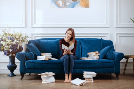 Téléchargez les photos : Jeune femme parmi des piles de livres sur un canapé spacieux dans le salon. - en image libre de droit