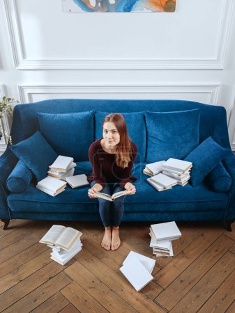Photo for Young surprised woman among stacks of books on a sofa in the living room. - Royalty Free Image