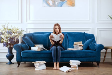 Téléchargez les photos : Jeune femme ennuyée avec un livre sur un canapé spacieux dans le salon. - en image libre de droit